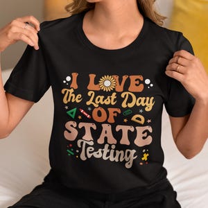 Può includere: T-shirt nera con la scritta "I Love The Last Day Of State Testing" in stile retrò. Il design include un girasole e grafiche scolastiche nei toni dell'arancione, marrone e verde.