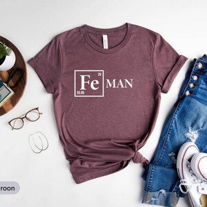 a casual outfit consisting of a maroon t-shirt with a white "Fe Man" text, a pair of blue jeans, white sneakers, and a pair of glasses. The outfit is arranged on a white background, with a potted plant and a camera visible in the background.