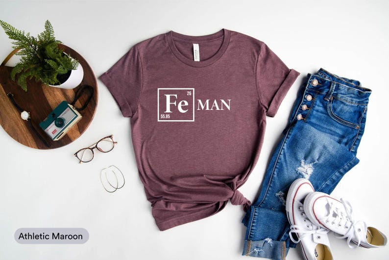 a casual outfit consisting of a maroon t-shirt with a white "Fe Man" text, a pair of blue jeans, white sneakers, and a pair of glasses. The outfit is arranged on a white background, with a potted plant and a camera visible in the background.