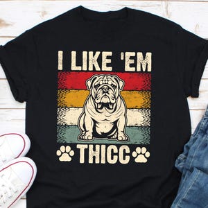 May include: Black t-shirt with a graphic of a bulldog and the text "I LIKE 'EM THICC". The design features a bulldog illustration in a retro color block design with red, yellow, and teal stripes. Paw prints flank the word "THICC".