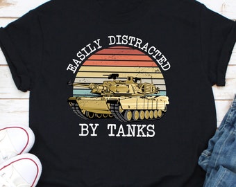 May include: Black t-shirt featuring a tan tank graphic with the text "EASILY DISTRACTED BY TANKS" in white. The design includes a retro sunset background. The shirt has rolled-up sleeves and a casual style.