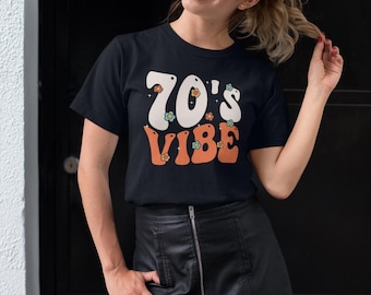 Puede incluir: Camiseta negra con el texto "70's Vibe" en fuente retro, con detalles blancos, naranjas y florales. La modelo lleva una falda de cuero negra. El tema general es la moda vintage.
