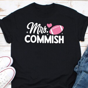 Puede incluir: Camiseta negra con texto blanco que dice "Mrs. Commish" y una pelota de fútbol rosa con un corazón rosa encima del texto.