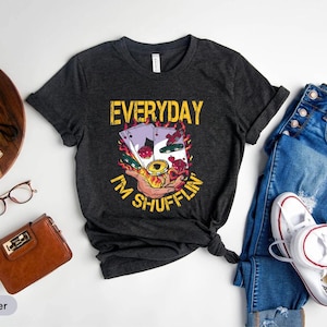 Puede incluir: Camiseta gris oscuro con el texto "EVERYDAY I'M SHUFFLIN'" en amarillo. El gráfico presenta naipes, llamas y una mano sosteniendo fichas de póquer. La camiseta se combina con vaqueros azules y zapatillas blancas.