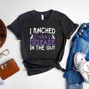 May include: Dark heather t-shirt with the text "I Punched Crohn's Disease in the Gut" in white and purple. The shirt is paired with blue jeans and white sneakers. A brown wallet, glasses, and a succulent plant are also in the image.
