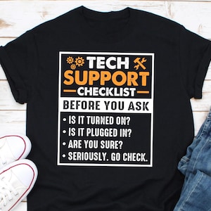 Può includere: T-shirt nero con la scritta "TECH SUPPORT CHECKLIST BEFORE YOU ASK". L'elenco di controllo include domande come "È ACCESO?" e "È COLLEGATO?" Il design presenta testo arancione e bianco.