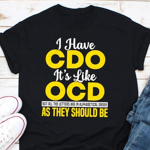 May include: Black t-shirt with the text "I Have CDO It's Like OCD But All The Letters Are In Alphabetical Order As They Should Be" in white and yellow. The shirt is folded, with white trainers and blue jeans visible.