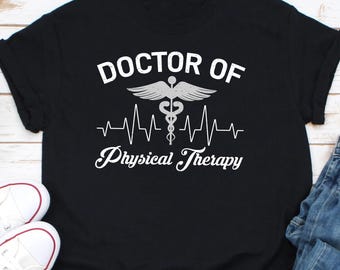 May include: Black t-shirt with white text and a medical symbol. The text reads "DOCTOR OF Physical Therapy" with a stylized heartbeat line. The shirt is folded, with white sneakers and blue jeans visible in the background.
