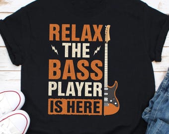 Puede incluir: Camiseta negra con el texto "Relax the Bass Player is Here" en naranja y un gráfico de guitarra eléctrica marrón.
