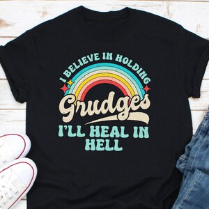 Op de afbeelding: Zwart t-shirt met een retro regenboogafbeelding en de tekst "I BELIEVE IN HOLDING GRUDGES I'LL HEAL IN HELL". Het ontwerp heeft een vintage lettertype en een regenboogboog. Het shirt wordt gecombineerd met witte sneakers en een blauwe jeans.