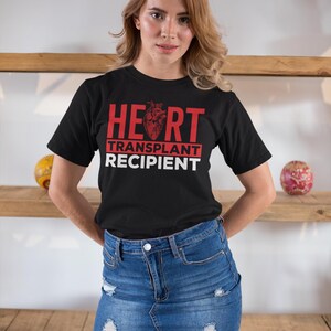 May include: A black t-shirt with red text that reads "HEART TRANSPLANT RECIPIENT". The text is in a bold font and the word "HEART" has a red anatomical heart graphic in the middle of the word. The person wearing the shirt is standing in front of a wooden shelf.