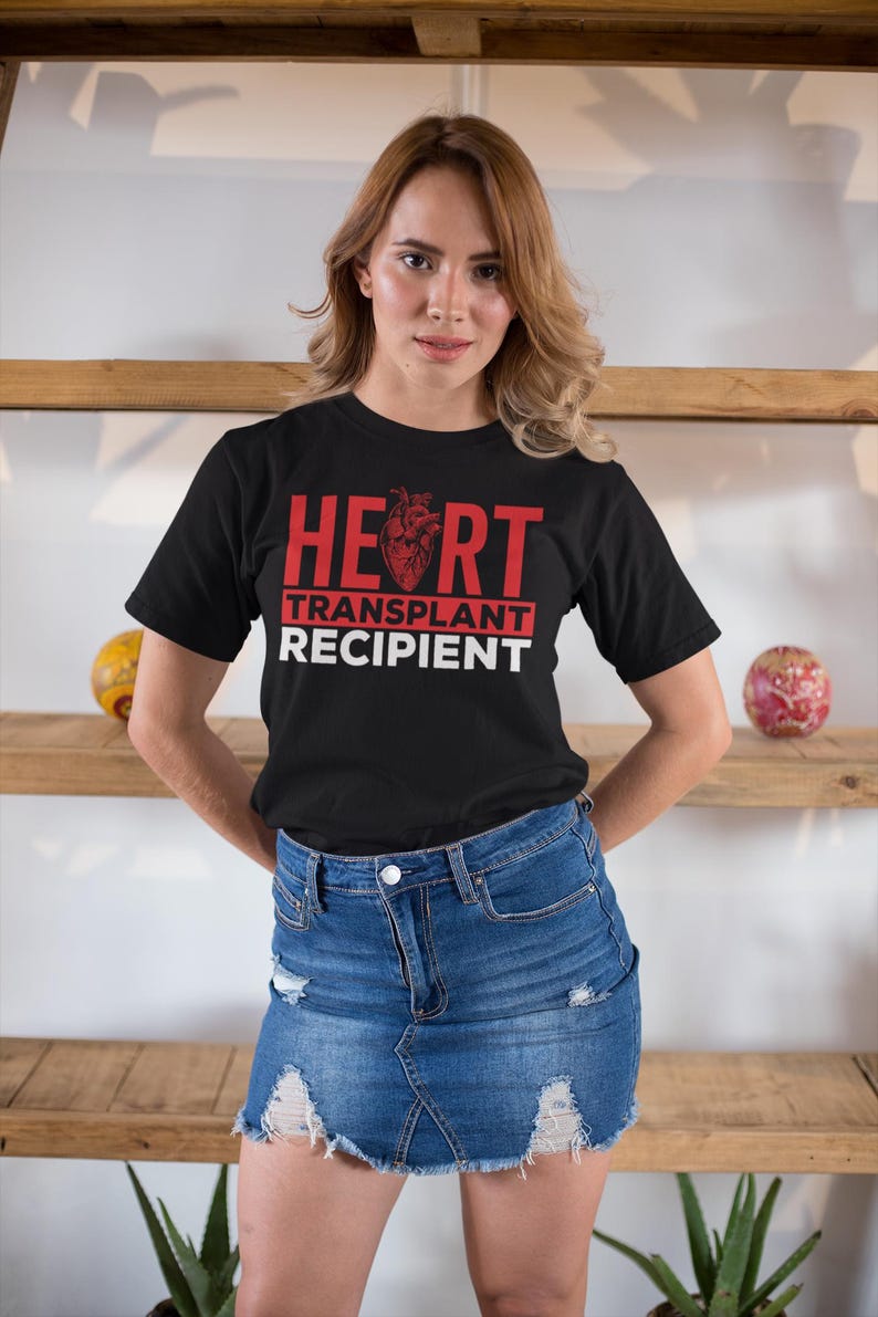 May include: A black t-shirt with red text that reads "HEART TRANSPLANT RECIPIENT". The text is in a bold font and the word "HEART" has a red anatomical heart graphic in the middle of the word. The person wearing the shirt is standing in front of a wooden shelf.