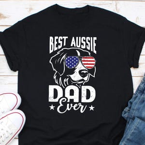 May include: Black t-shirt featuring a white graphic of an Australian Shepherd wearing sunglasses with an American flag design. The text "Best Aussie Dad Ever" is printed below the dog.