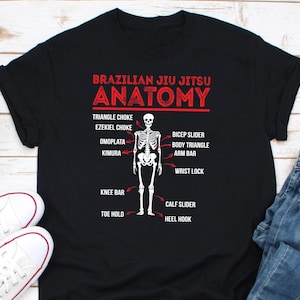 May include: Black t-shirt featuring a white skeleton illustration and red text that reads "Brazilian Jiu Jitsu Anatomy". The skeleton is labelled with various Jiu Jitsu techniques, including Triangle Choke, Ezekiel Choke, Omoplata, Kimura, Bicep Slider, Body Triangle, Arm Bar, Wrist Lock, Knee Bar, Calf Slider, Toe Hold, and Heel Hook.