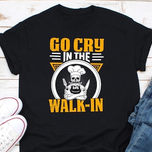 Könnte beinhalten: Schwarzes T-Shirt mit einer gelben Grafik eines Kochs mit einem Totenkopfgesicht, der ein Messer und eine Gabel hält. Der Text "Go Cry In The Walk-In" ist in Gelb über dem Koch geschrieben.