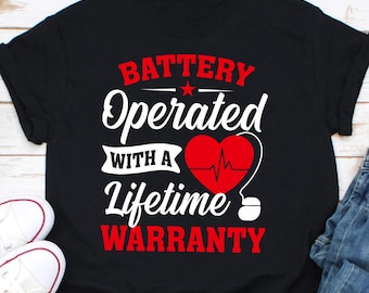 May include: Black t-shirt with the text "BATTERY Operated WITH A Lifetime WARRANTY" in red and white. A red heart with a heartbeat line is in the center of the design. White sneakers are visible in the lower left corner.