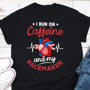 Puede incluir: Camiseta negra con un corazón rojo y un gráfico de ECG. El texto "I run on Caffeine and my Pacemaker" está impreso en la camiseta.