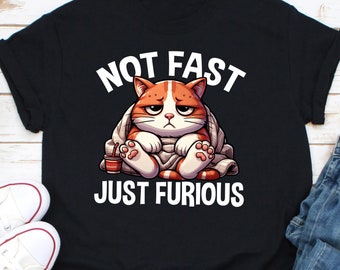 May include: Black t-shirt with the text "NOT FAST JUST FURIOUS" above and below a cartoon illustration of a grumpy orange and white cat wrapped in a blanket. The cat is holding a small cup.