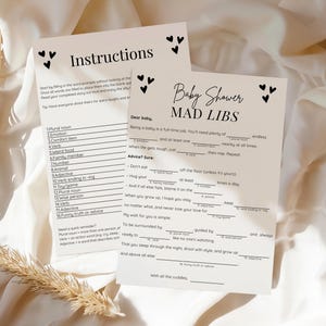 May include: Two white paper sheets with black text and heart illustrations. One sheet reads "Instructions" and the other is a "Baby Shower MAD LIBS" game. The sheets are on a white fabric background with a dried plant stem.
