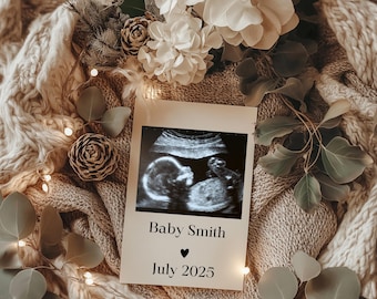 Pode incluir: Um cartão branco com uma imagem de ultrassom preta de um bebê. O cartão tem o texto "Baby Smith" e "Julho 2025" com um símbolo de coração preto entre as duas linhas de texto. O cartão está sobre um cobertor de malha marrom com luzes brancas e folhas verdes.