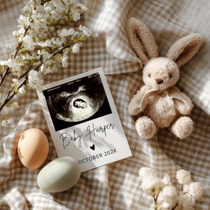 May include: A pregnancy announcement card with a sonogram image, the text "Baby Harper", and "OCTOBER 202X". A plush bunny, two eggs, and white flowers are arranged on a tan and white checkered fabric.