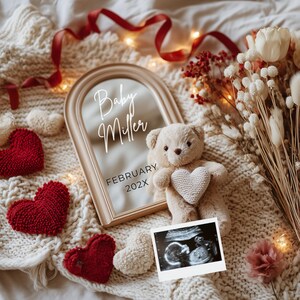 May include: A cream-coloured teddy bear holding a heart, beside a framed mirror with the text "Baby Miller FEBRUARY 202X". Red and white heart-shaped decorations and a sonogram are also present. The scene is set on a cream-coloured knitted blanket.