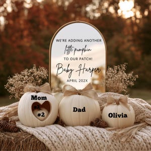 以下が含まれることがあります： Mom、Dad、Oliviaと書かれたベージュの麻のリボン付きの白い装飾用カボチャ3個。Momのカボチャにはハート型の切り抜きがあり、中に#2と書かれた小さなカボチャが入っている。アーチ型の鏡には「WE'RE ADDING ANOTHER little pumpkin TO OUR PATCH! Baby Harper APRIL 202X」という文字が表示されている。