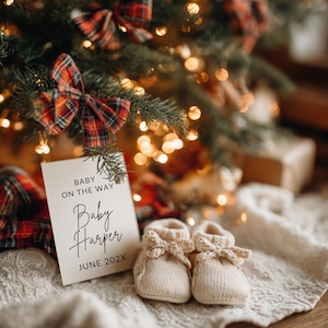 May include: A pregnancy announcement card with the text "Baby on the Way" and "Baby Harper June 202X" sits in front of a Christmas tree. Two small, cream-colored knitted baby booties are in front of the card. The scene is festive with soft lighting.