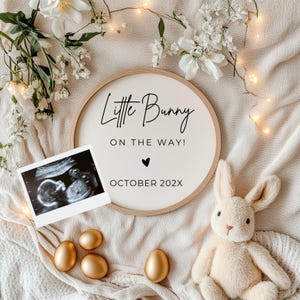May include: A white wooden round sign with the text "Little Bunny On The Way! October 202X" in black lettering. A black heart is above the date. A black and white ultrasound image is in the foreground. There are white flowers and a stuffed bunny in the background.