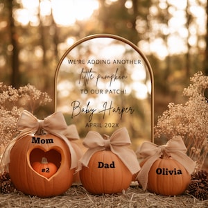 May include: Three orange pumpkins with tan burlap bows. The largest pumpkin has a heart-shaped cutout containing a small pumpkin labelled #2 and is marked Mom. The others are labelled Dad and Olivia. An arched sign reads: WE'RE ADDING ANOTHER little pumpkin TO OUR PATCH! Baby Harper APRIL 202X.