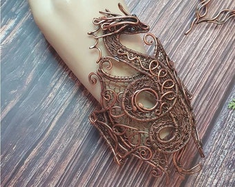 Puede incluir: Un brazalete de dragón de alambre de cobre con detalles intrincados. El brazalete está hecho de alambre que se teje para crear un diseño de dragón. La cabeza del dragón está mirando hacia adelante y sus alas están extendidas. El brazalete es una hermosa pieza de joyería que sería un gran regalo para cualquier amante de los dragones.