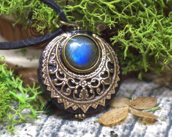 Puede incluir: Un colgante de latón con una piedra preciosa de labradorita azul engastada en un diseño de media luna. El colgante está rodeado de intrincados detalles florales y cuelga de un cordón de cuero negro.