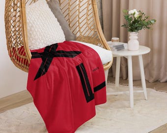 Può includere: Una coperta rossa con disegni geometrici neri e la parola "HUGS" in bianco. La coperta è drappeggiata su una sedia in vimini.