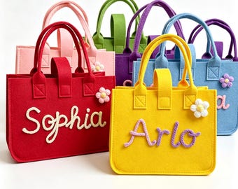 May include: A collection of rectangular felt tote bags in various colors including red, yellow, blue, pink, green, and purple. Each bag features two handles, a small crocheted flower accent, and a personalized name written in cursive rope text.