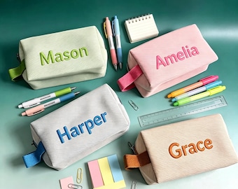 May include: Four rectangular pencil cases in light green, pink, gray, and beige, each featuring a textured surface and a side handle. The names Mason, Amelia, Harper, and Grace are embroidered on the front. Pens, a ruler, and paper clips are included.