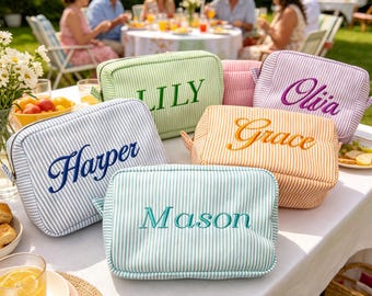 May include: A collection of five rectangular zippered pouches made of striped fabric in light green, light blue, orange, pink, and lavender. Each pouch features a name embroidered in a contrasting thread color: Lily, Harper, Mason, Grace, and Olivia.
