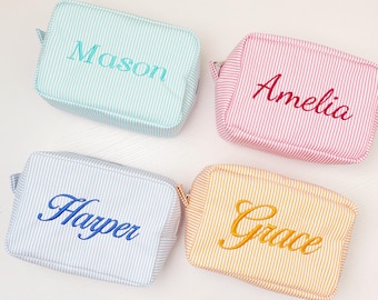 May include: Four rectangular zippered pouches with a white pinstripe pattern. The top left pouch is light blue with the name Mason embroidered in teal. The top right is pink with Amelia in dark pink. Bottom left is light blue with Harper in blue. Bottom right is yellow with Grace in gold.