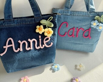 May include: Two denim tote bags with top handles. The dark blue bag features the name Annie in pink yarn and two yellow crocheted flowers. The light blue bag features the name Cara in pink yarn and two blue crocheted flowers. Loose flowers are scattered.