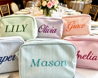 May include: A collection of striped cosmetic bags in various pastel colors, each embroidered with a different name. The bags are arranged on a white surface, with a blurred background of a dining setting, including a floral centerpiece.