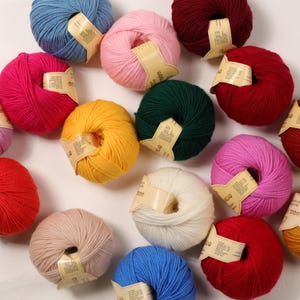 Op de afbeelding: Assortiment van kleurrijke garenbollen in verschillende tinten roze, rood, blauw, geel, groen, wit en beige. Elke bol heeft een label met het woord "Wol" erop gedrukt.