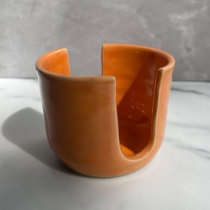 May include: A bright orange ceramic yarn bowl with a smooth, glossy finish. The bowl features a rounded base and a wide, U-shaped cutout on one side, designed to hold a ball of yarn while allowing the strand to feed through the opening smoothly.