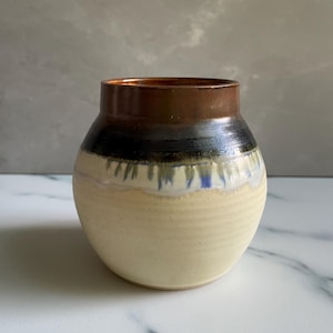May include: A ceramic vase with a rounded body and a wide opening. The upper rim is a metallic bronze, transitioning into a dark black band that drips into a light cream-colored base. The surface features subtle horizontal ridges along the lower half.