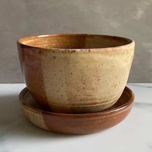 May include: A ceramic bowl and matching saucer set featuring a two-tone glaze. The upper portion of the bowl and the outer rim of the saucer are finished in a speckled beige, while the lower section and base are a solid, deep reddish-brown.