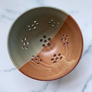 May include: A handmade ceramic colander with a two-tone design. One half is a muted green, the other a warm brown. The bowl has numerous small holes for draining and a rustic, artisanal aesthetic.