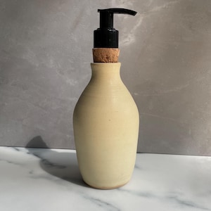 May include: A cream-colored ceramic soap dispenser with a smooth, matte finish and a rounded body. The top features a natural cork stopper fitted with a black plastic pump mechanism for dispensing liquid soap or lotion in a functional design.