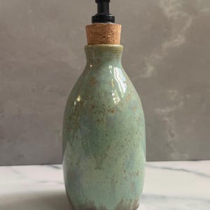 May include: A ceramic soap dispenser with a green and brown glaze. The dispenser has a black pump and a cork stopper. The bottle is approximately 7 inches tall.