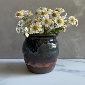 May include: A small, rounded ceramic vase with a glossy glaze featuring blended tones of deep blue, dark green, and earthy brown. The vase is filled with a bouquet of white daisies with bright yellow centers, showcasing delicate petals and green stems.