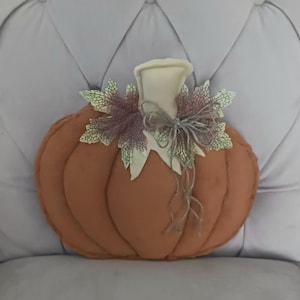 May include: A plush, orange pumpkin decoration with a cream-colored stem and decorative leaves. The leaves are a mix of green and purple, and the stem is tied with twine. This fall-themed item is perfect for seasonal home decor.