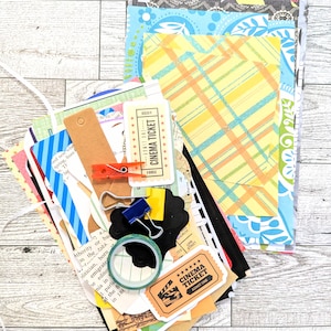Puede incluir: Una colección de recortes de papel de colores, que incluyen un papel a rayas azul y blanco, un papel a cuadros amarillo y naranja y un papel con estampado en blanco y negro. También hay varios billetes de papel con el texto "CINEMA TICKET" y un rollo de cinta washi.