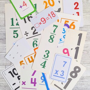 May include: A collection of vintage flashcards with arithmetic problems. The cards feature numbers and equations in various colors, including green, orange, purple, and blue. The cards are scattered on a light-colored wooden surface.